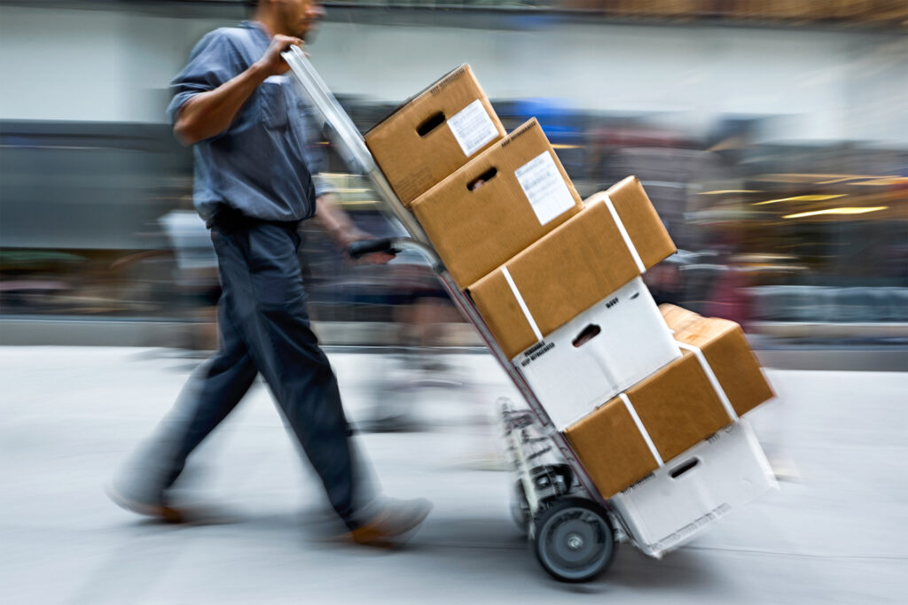 A delivery professional pushing a handcart loaded with shipping boxes against a blurred background