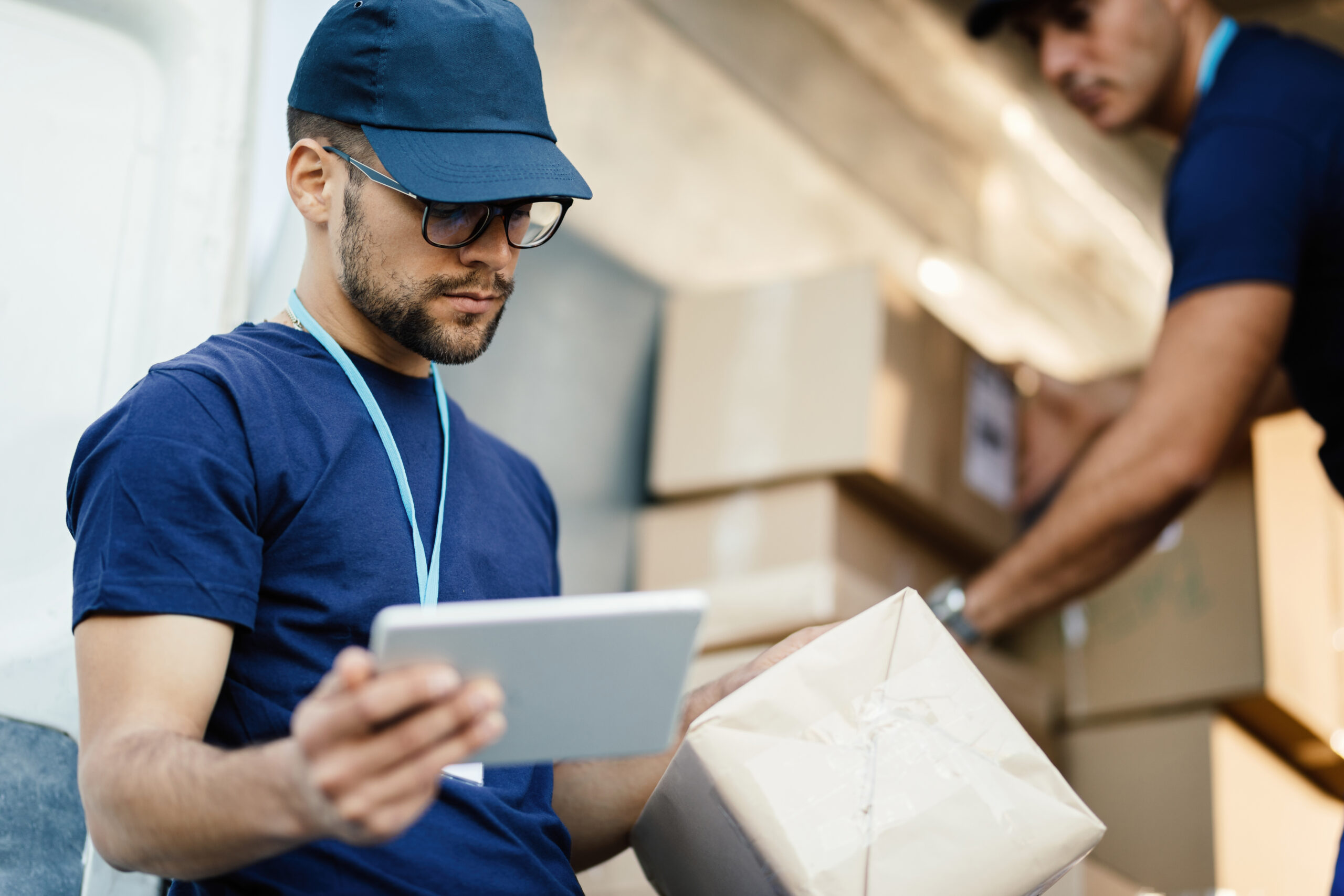 A 3PL professional using a tablet to check a package while another professional handles boxes in a delivery vehicle.