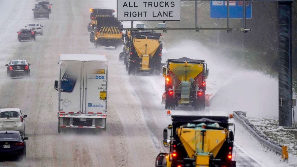 Logistics trucks and cars navigating an icy, snow-covered highway as snow plows clear the road ahead, illustrating the impact of adverse weather on transportation logistics. A sign directs all trucks to the right lane.