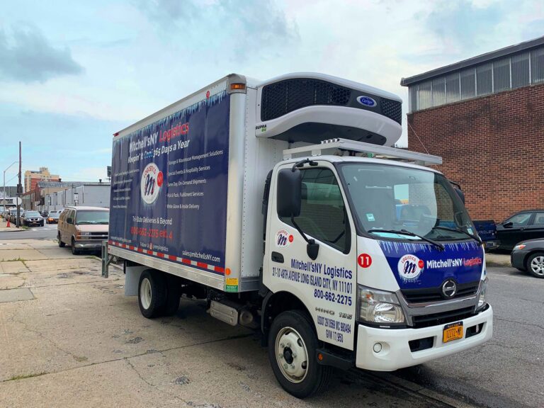 A Mitchell’sNY Logistics refrigerated delivery truck parked on a city street, displaying the company’s branding and contact information.