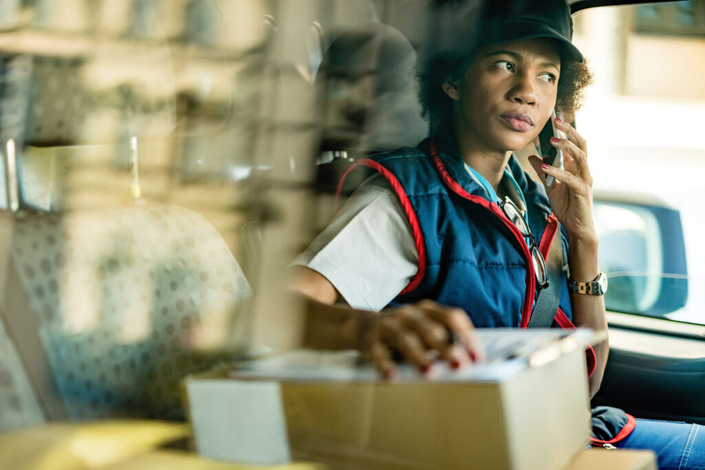 A Mitchell’sNY Logistics delivery professional sitting in a van, holding a package and talking on a mobile phone, viewed through the vehicle window.