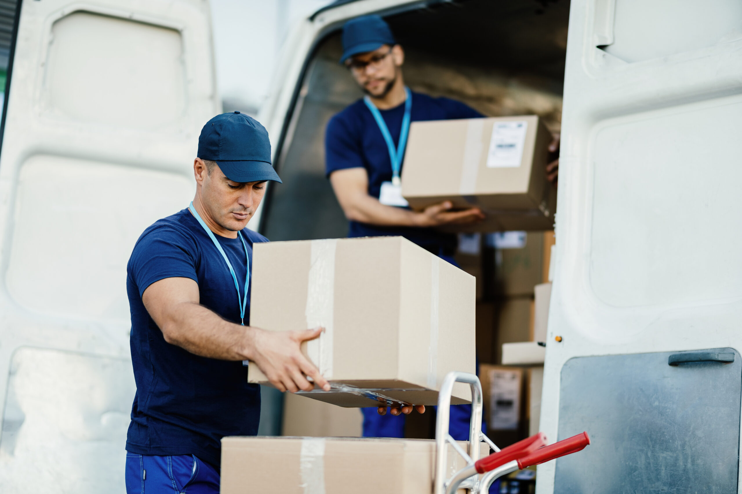 Two Mitchell’sNY Logistics delivery professionals in blue uniforms unload cardboard boxes from a white van, preparing packages for distribution.