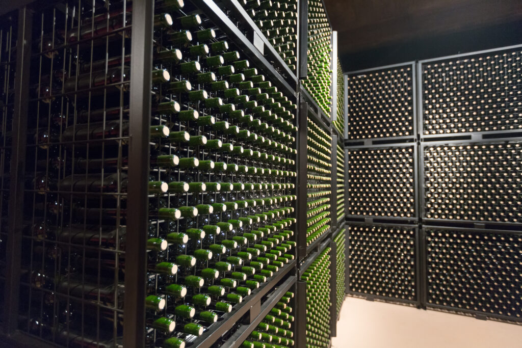 A large wine cellar filled with hundreds of green glass wine bottles neatly stored horizontally in black metal racks that cover the walls from floor to ceiling, creating an organized and visually striking grid pattern.