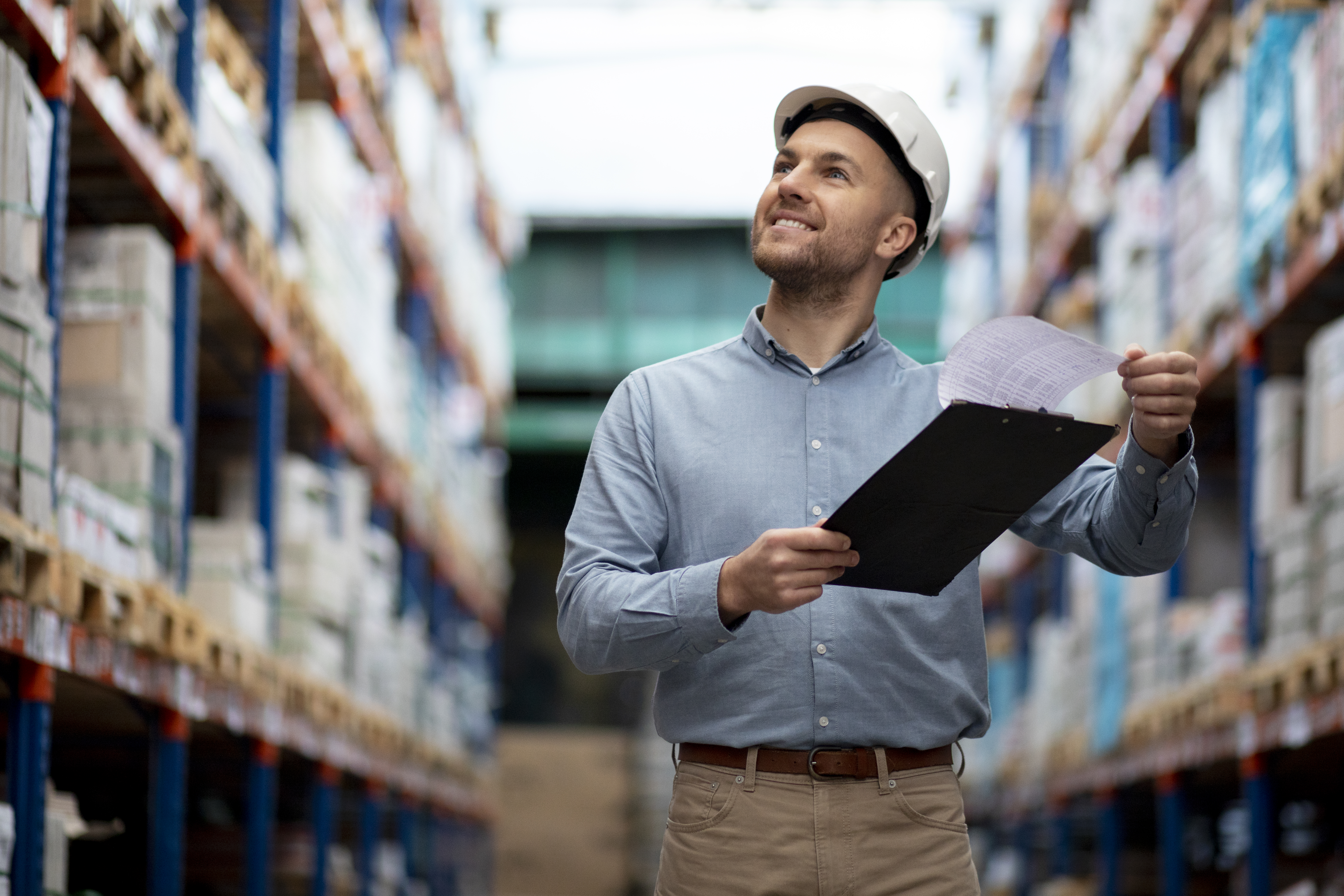 A warehouse professional wearing a hardhat and carrying a clipboard checking the inventory.