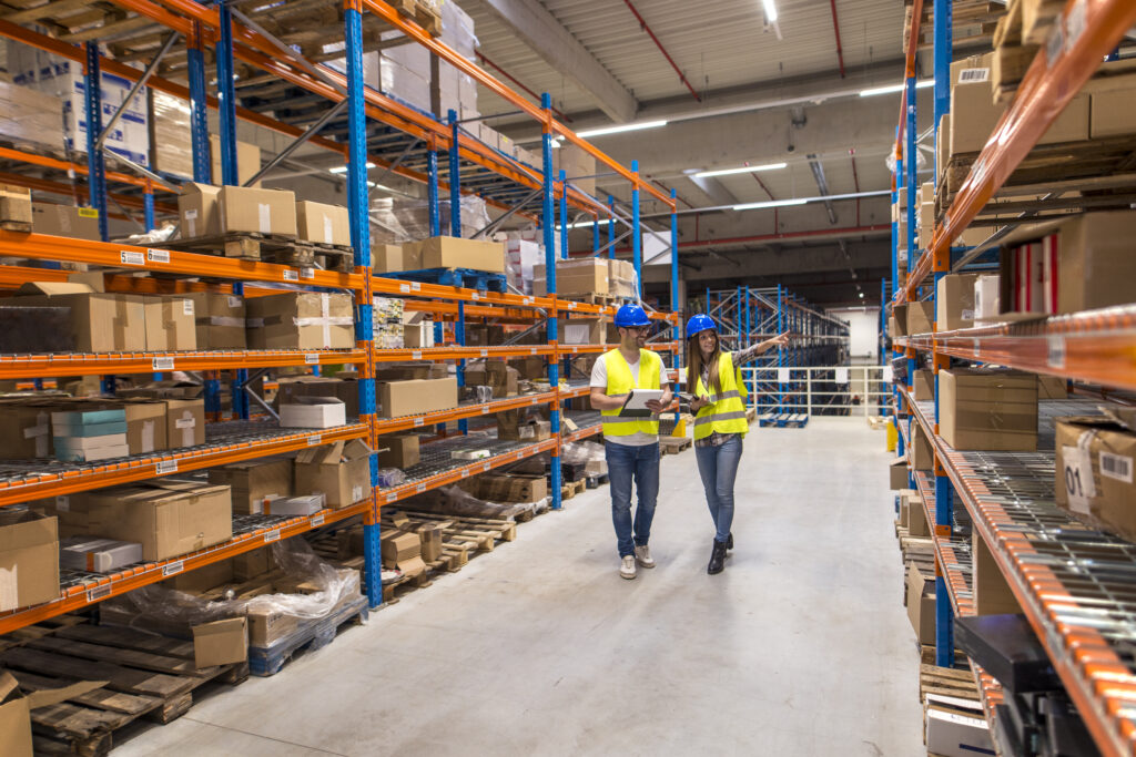 Two warehouse workers walking in distribution storage area discussing about logistics and organization.