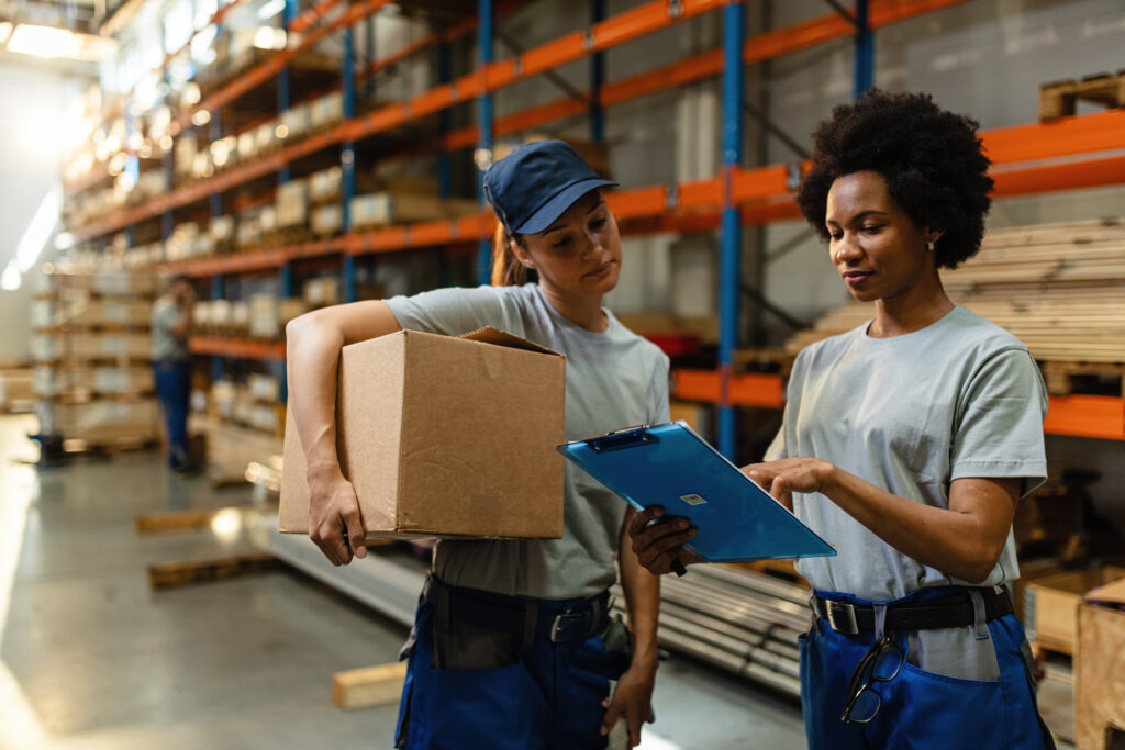 Two warehouse workers review a shipment checklist on a clipboard while one holds a cardboard box in a warehouse aisle with stocked shelving.