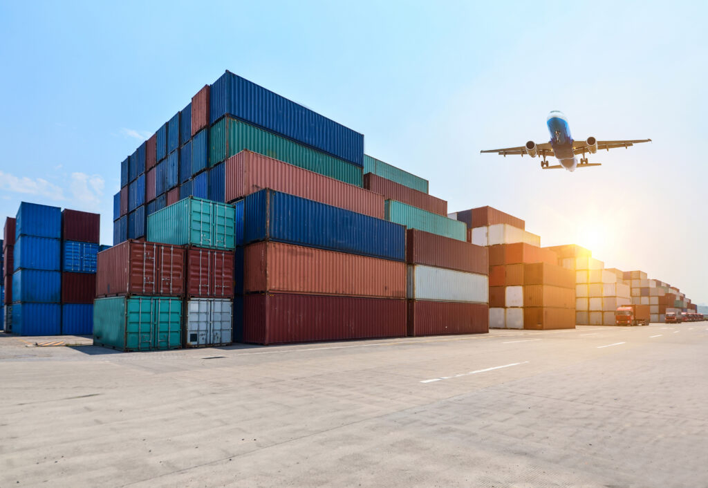 Wide view of stacked shipping containers at an industrial port yard, with a cargo plane flying overhead and trucks lined up in the distance at sunset.