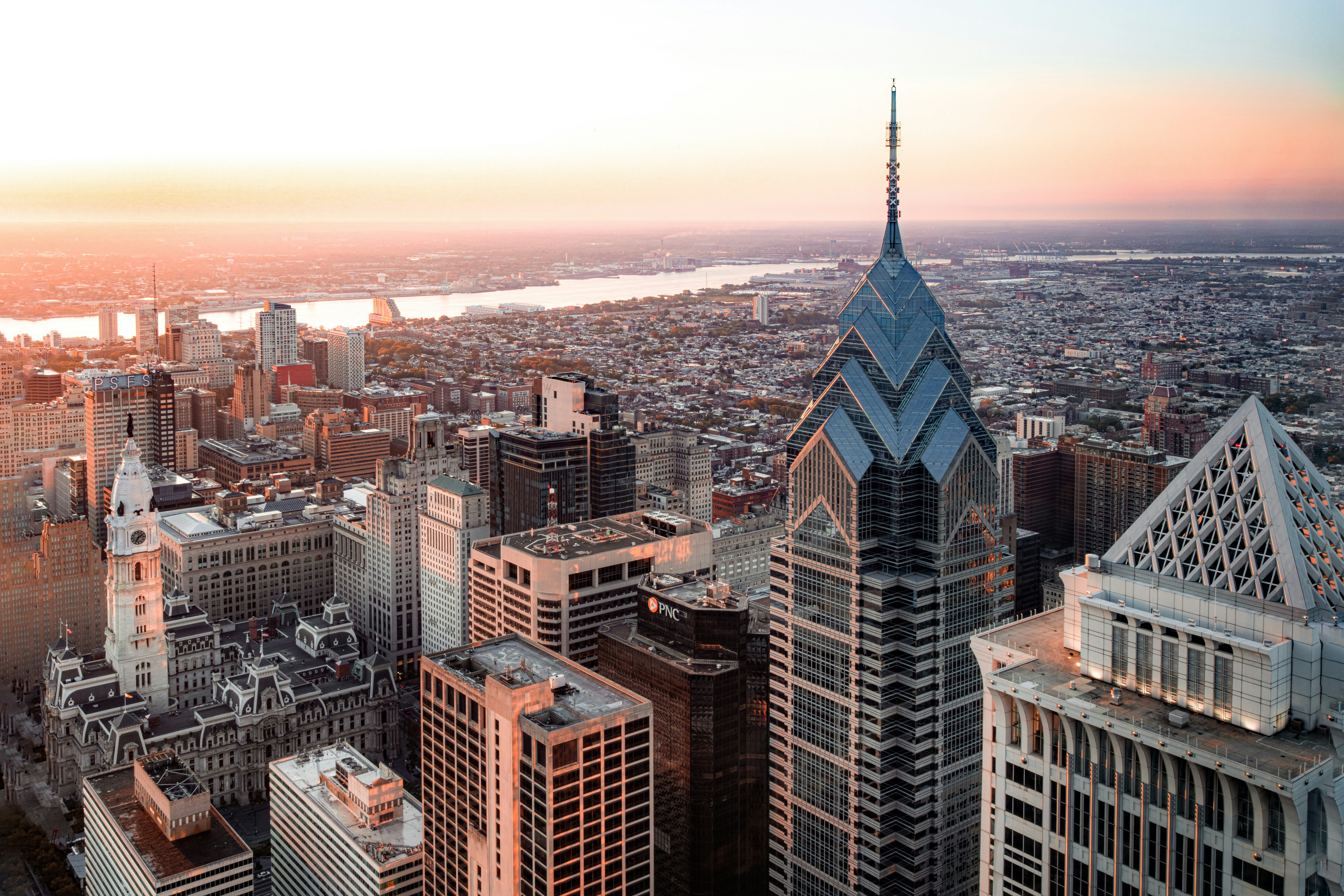 Aerial view of Center City Philadelphia skyline at sunset, featuring One Liberty Place, City Hall, and the Delaware River in the distance.