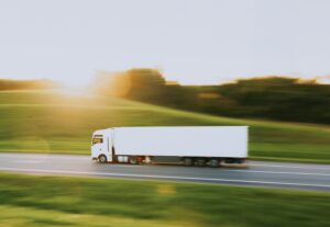 White semi-truck traveling along a highway through green countryside, with motion blur suggesting speed and regional transportation.